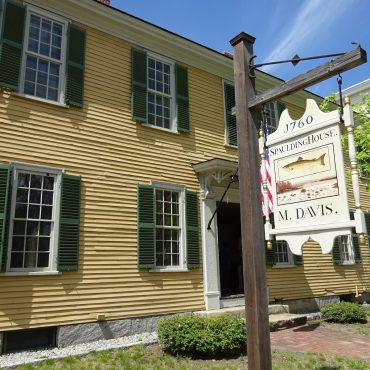 A mustard yellow house with dark green shutters and a white door, a wooden pole holds a white sign that reads 1760 Spaulding House M. Davis." In the center of the sign is a fish and a river bank