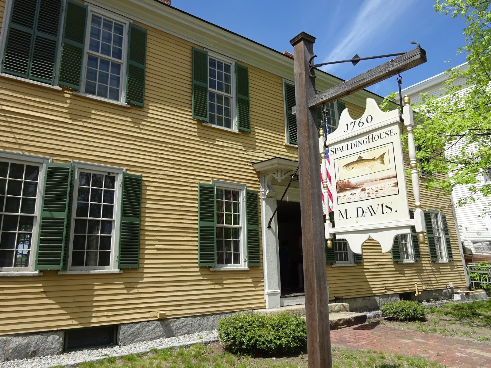 A mustard yellow house with dark green shutters and a white door, a wooden pole holds a white sign that reads 1760 Spaulding House M. Davis." In the center of the sign is a fish and a river bank