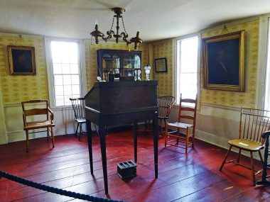 a standing desk with a pen, oil lamp, and hourglass, on the yellow wallpaper are portraits of various people, a degree from Dartmouth College to Dr. Joel Spalding, and a book case