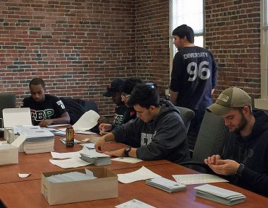 7 college fraternity students, stamp and help mail letters sitting at a conference table, a woman leaning over table helping nearby
