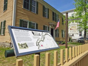 a mustard yellow house with green shutters and an open door, a yellow fence surrounds the house, a sign that reads "Villages at the Falls", with a map