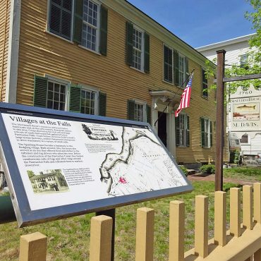 a mustard yellow house with green shutters and an open door, a yellow fence surrounds the house, a sign that reads "Villages at the Falls", with a map