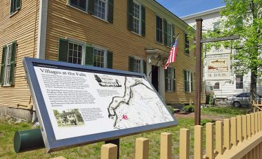 a mustard yellow house with green shutters and an open door, a yellow fence surrounds the house, a sign that reads "Villages at the Falls", with a map
