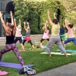 adults stand on yoga matts outside on the grass, and outdoor stage in front of them, their backs are to us with their arms in the air as the lunge foward