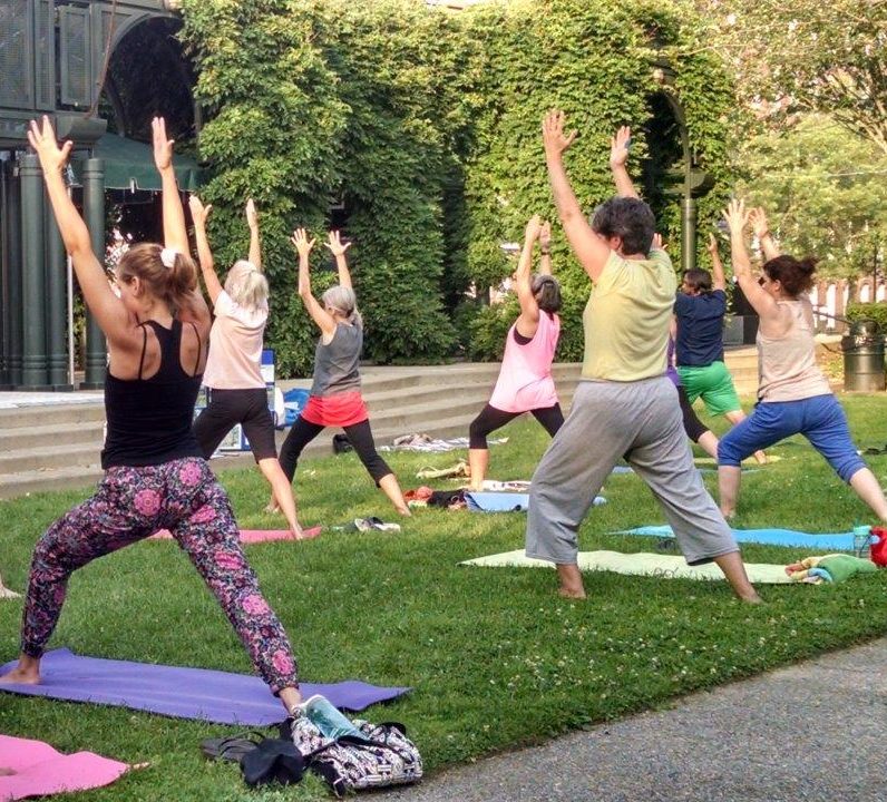 adults stand on yoga matts outside on the grass, and outdoor stage in front of them, their backs are to us with their arms in the air as the lunge foward