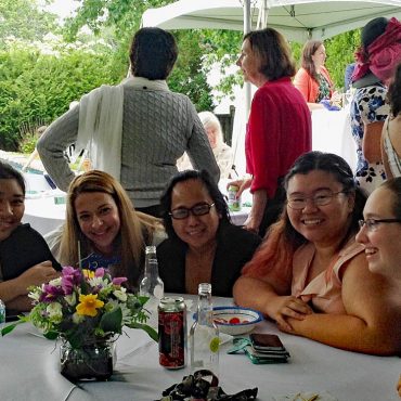 5 students sit a table with a floral centerpiece, they are smiling, adults mingl in the background of the outdoor event