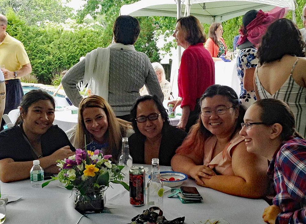 5 students sit a table with a floral centerpiece, they are smiling, adults mingl in the background of the outdoor event