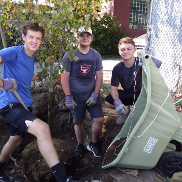 3 teens standing in a hole pouring dirt from a wheelbarrow, planting a new tree