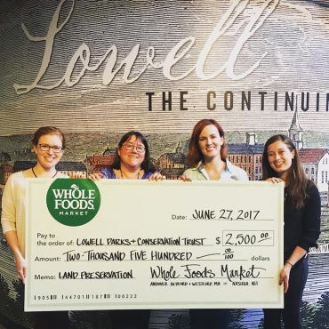 4 woman hold up a large check from Whole Foods Market, in front of a Lowell banner