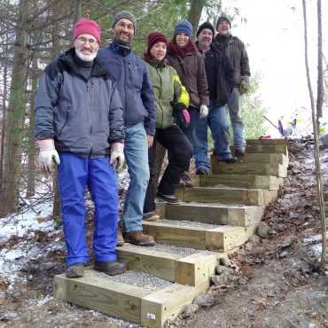 six people stand on a outdoor wooden stair case, pine trees behind them, dirt and snow on ground, they are in winter clothing