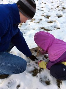 a man and child crouch on the snow examining an animal track, the man is pointing to the track adn the young girl is holding an ID brochure