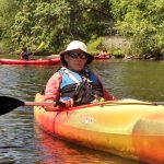 man with sunglasses and bucket hat in a canoe with 3 canoers in the background, green bushes and trees on river bank