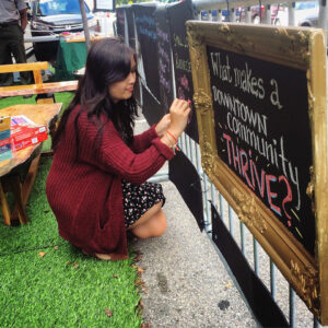 a woman kneels writing on a chalkboard that is hung on an outdoor fence - What makes a downtown community thrive?"