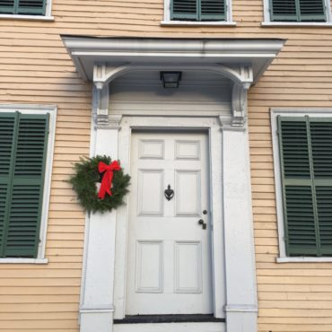 Historic mustard yellow house, dark green shutters, white door with a holiday wreath and red bow hanging to the side of the door