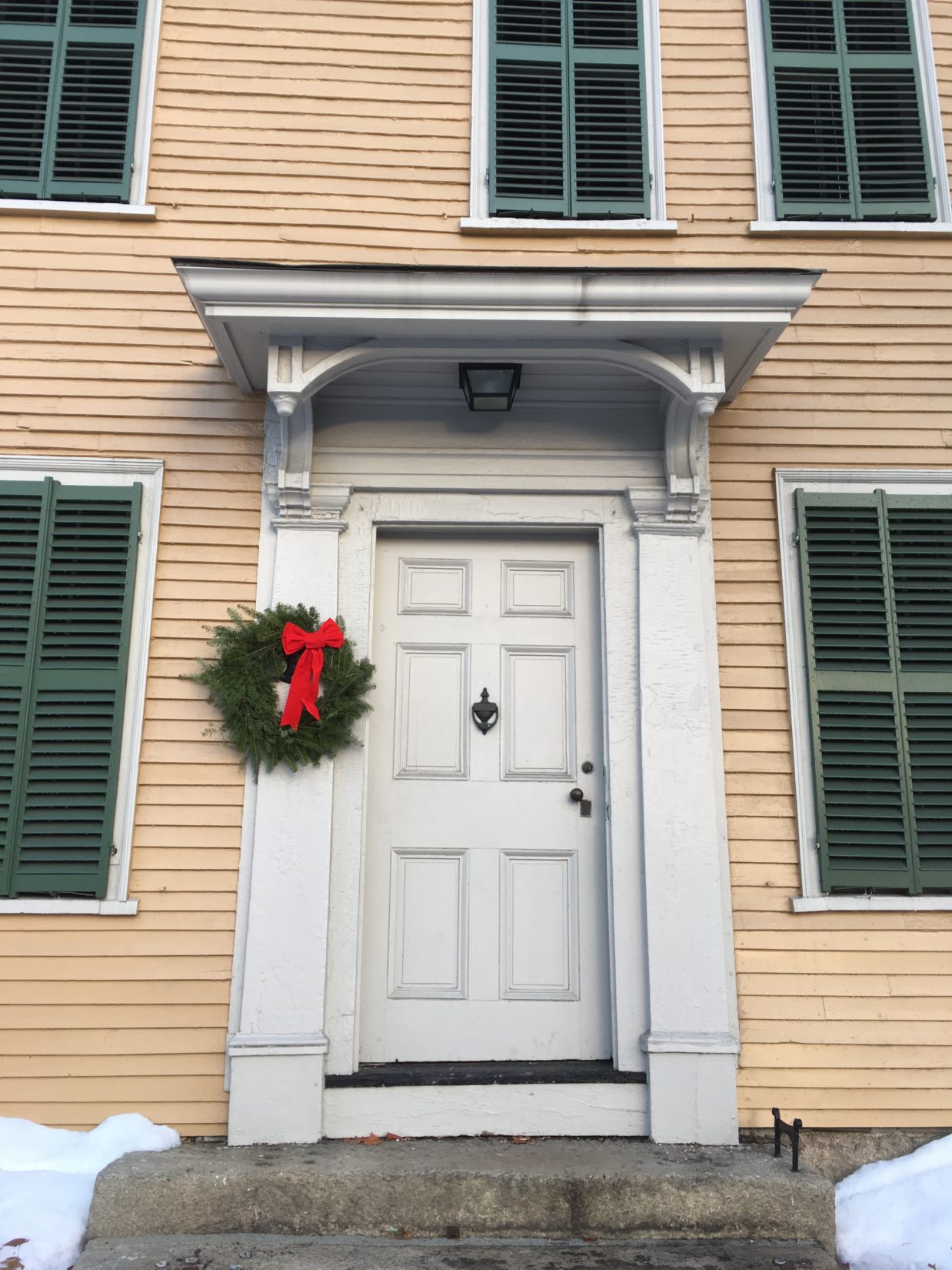 Historic mustard yellow house, dark green shutters, white door with a holiday wreath and red bow hanging to the side of the door