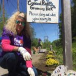 Fed Ex employee in purple smiles as she plants flowers under the Concord River Greenway Park sign, sign reads "Concord River Greenway Park Please carry out what you carry in"