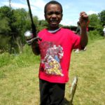 young boy holds up a fishing pole and caught fish by the river