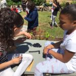 two girls on a stone wall play dominos as more children play on grass behind them